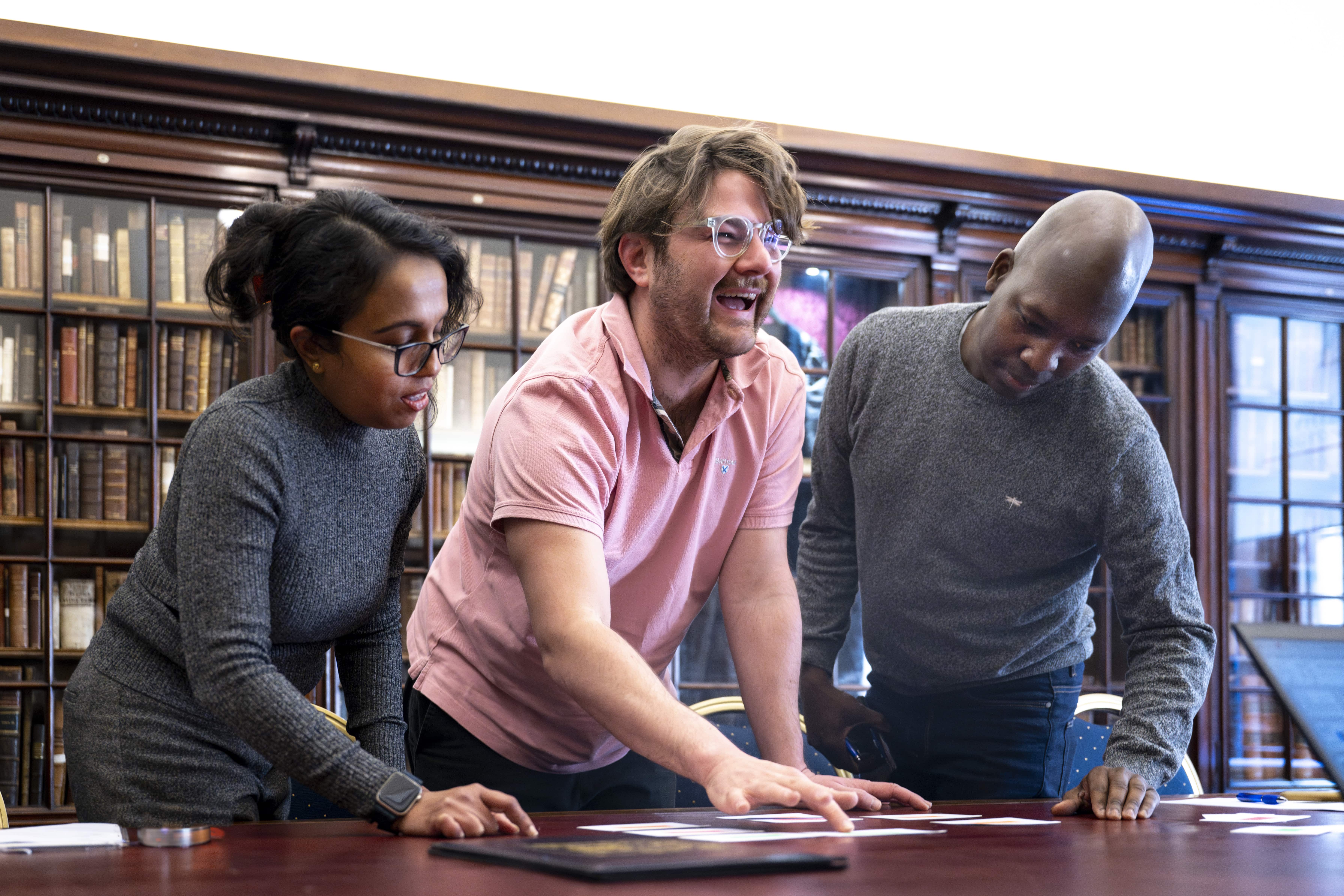 Three colleagues engage in a collaborative exercise around a table in a library setting, smiling and interacting with printed materials
