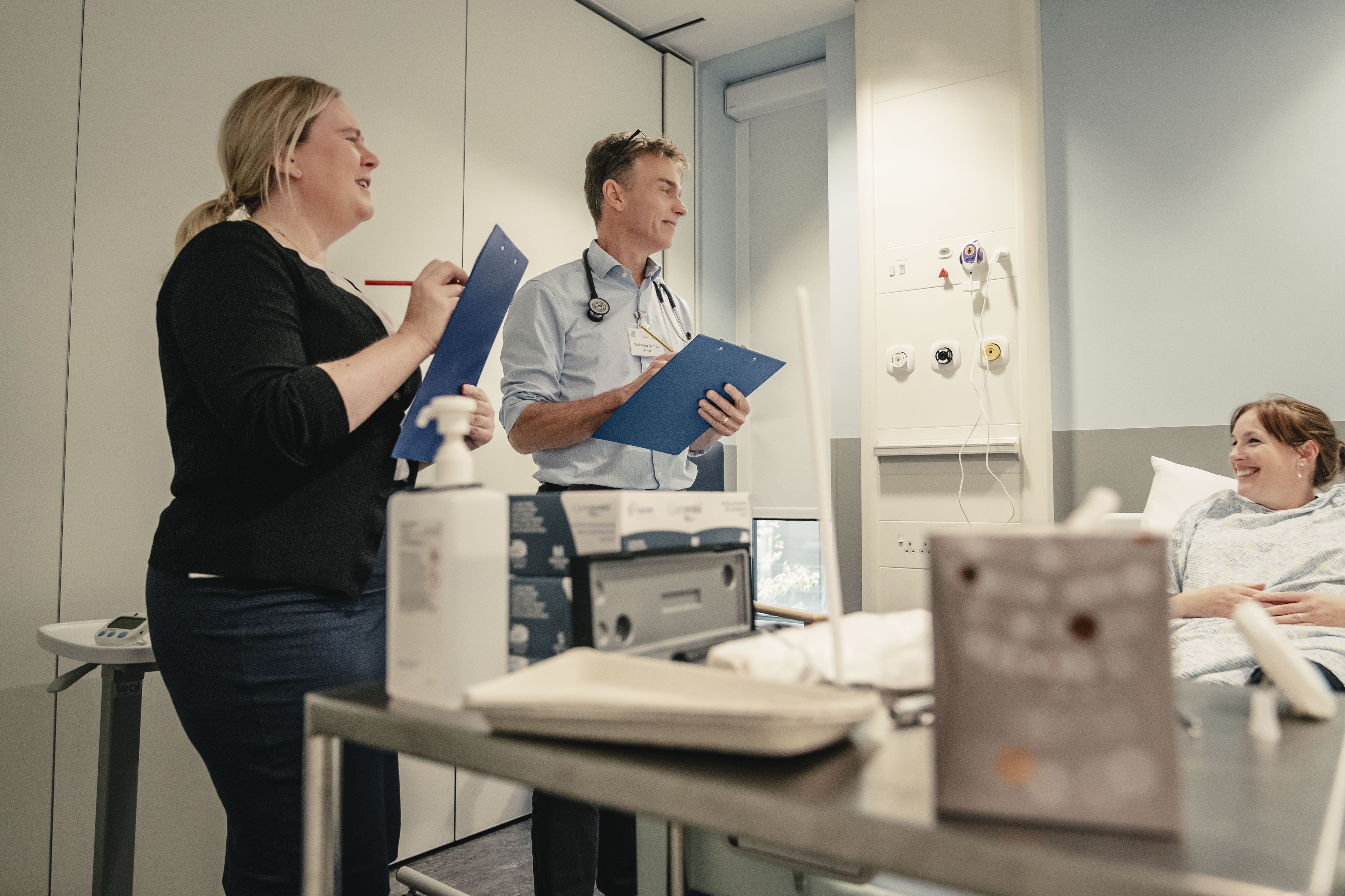 Two clinicians with clipboards observe and interact with a smiling patient in a hospital room during a training or assessment session