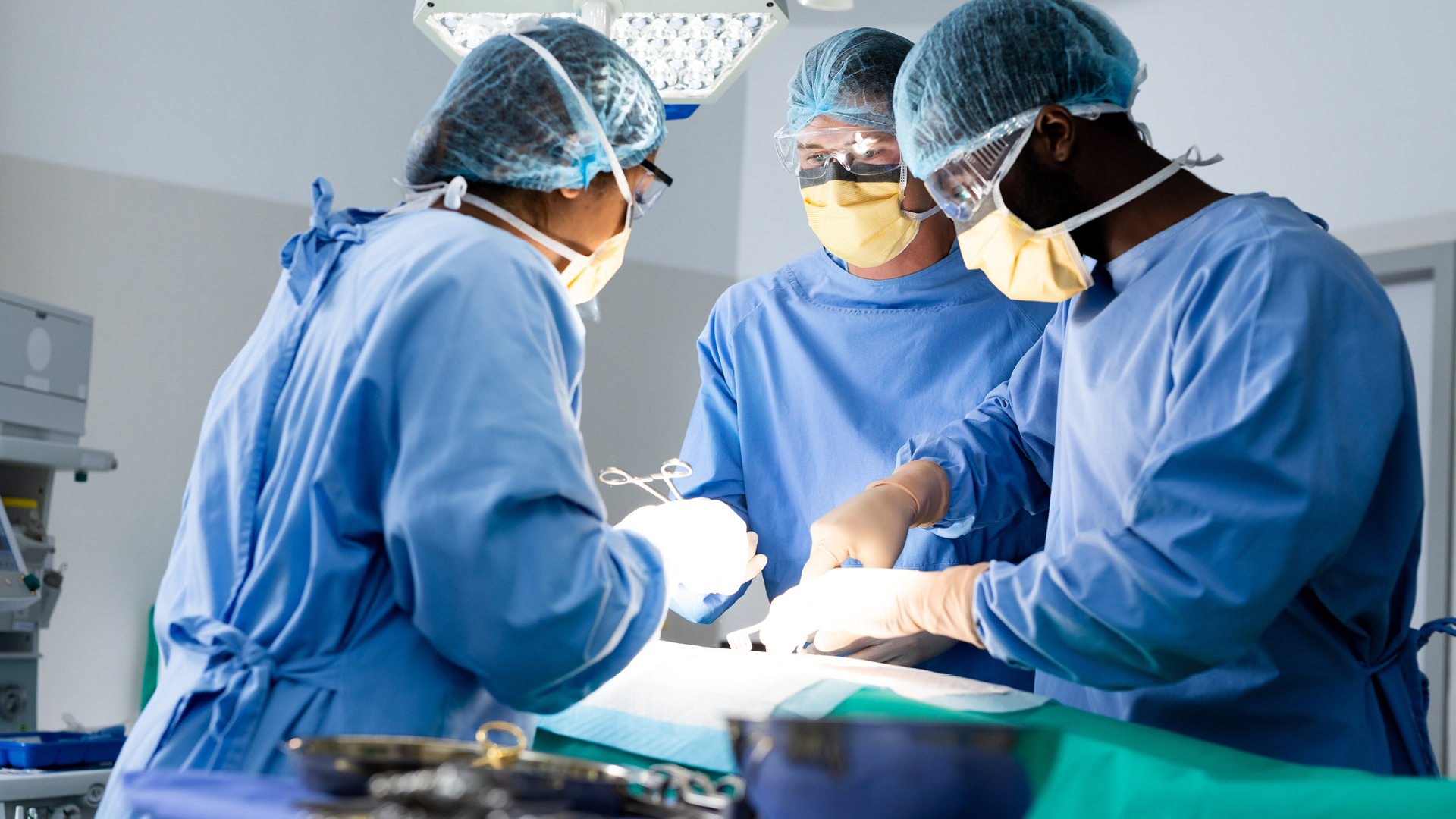 A group of surgeons in full sterile gown and masks perform a procedure under bright operating theatre lights