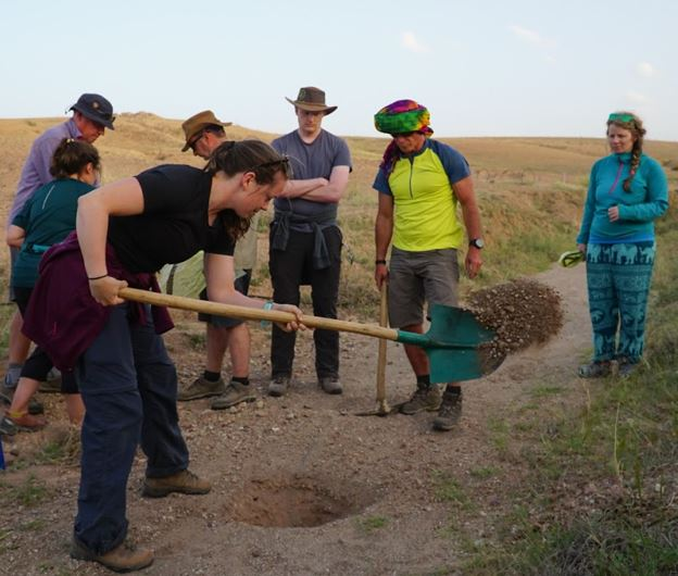A group of people in outdoor clothing observe as one participant shovels gravel into a hole on a dirt path in a rural, hilly landscape